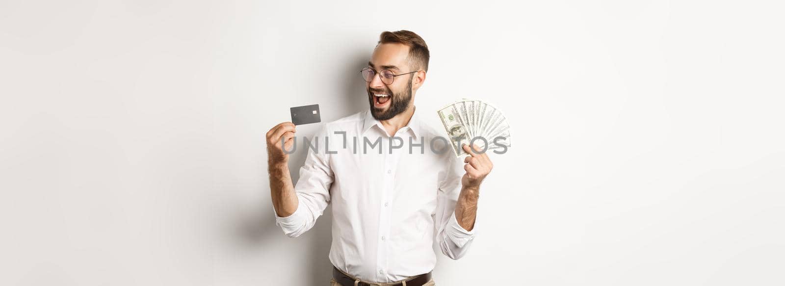 Excited businessman holding money and looking at credit card, standing over white background.