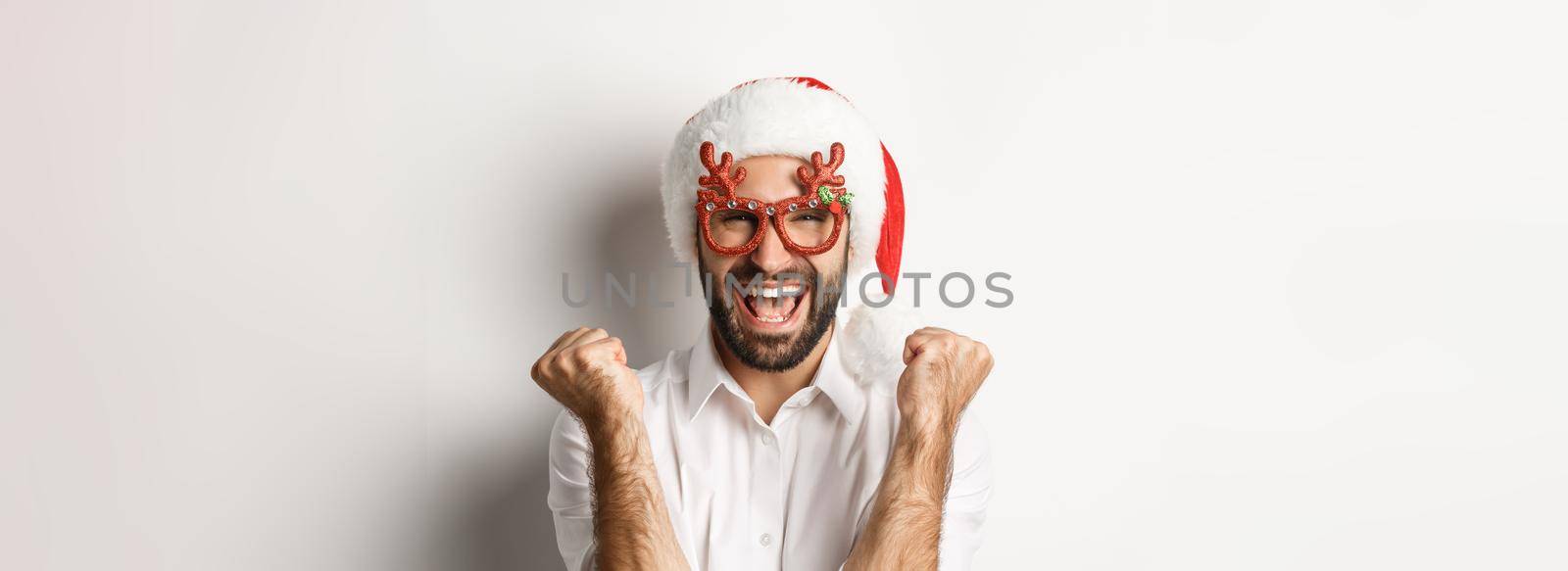 Close-up of man celebrating christmas or new year, wearing xmas party glasses and santa hat, rejoicing and shouting of joy, standing over white background by Benzoix