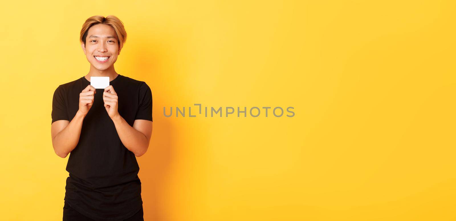 Excited happy asian guy smiling and showing credit card, standing yellow background by Benzoix