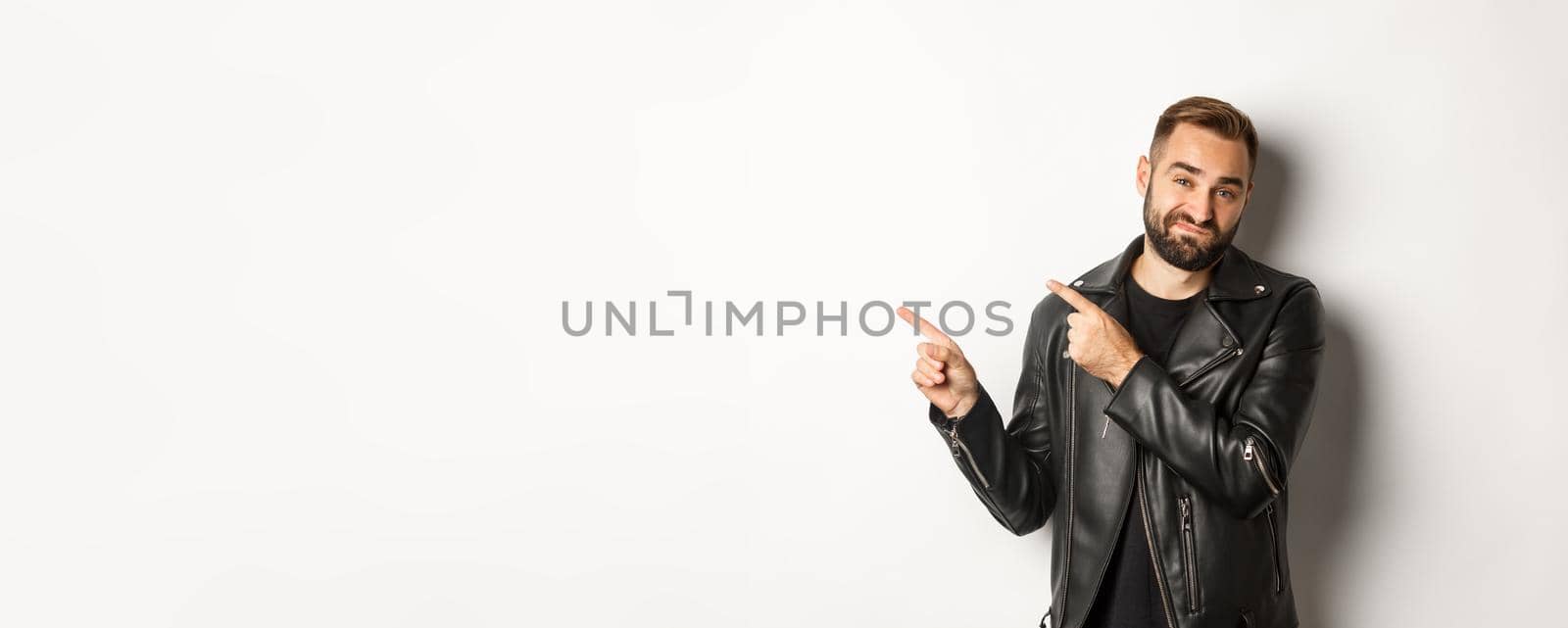 Skeptical and disapleased man in leather jacket, pointing fingers at upper left corner, showing bad promo offer, standing over white background reluctant by Benzoix