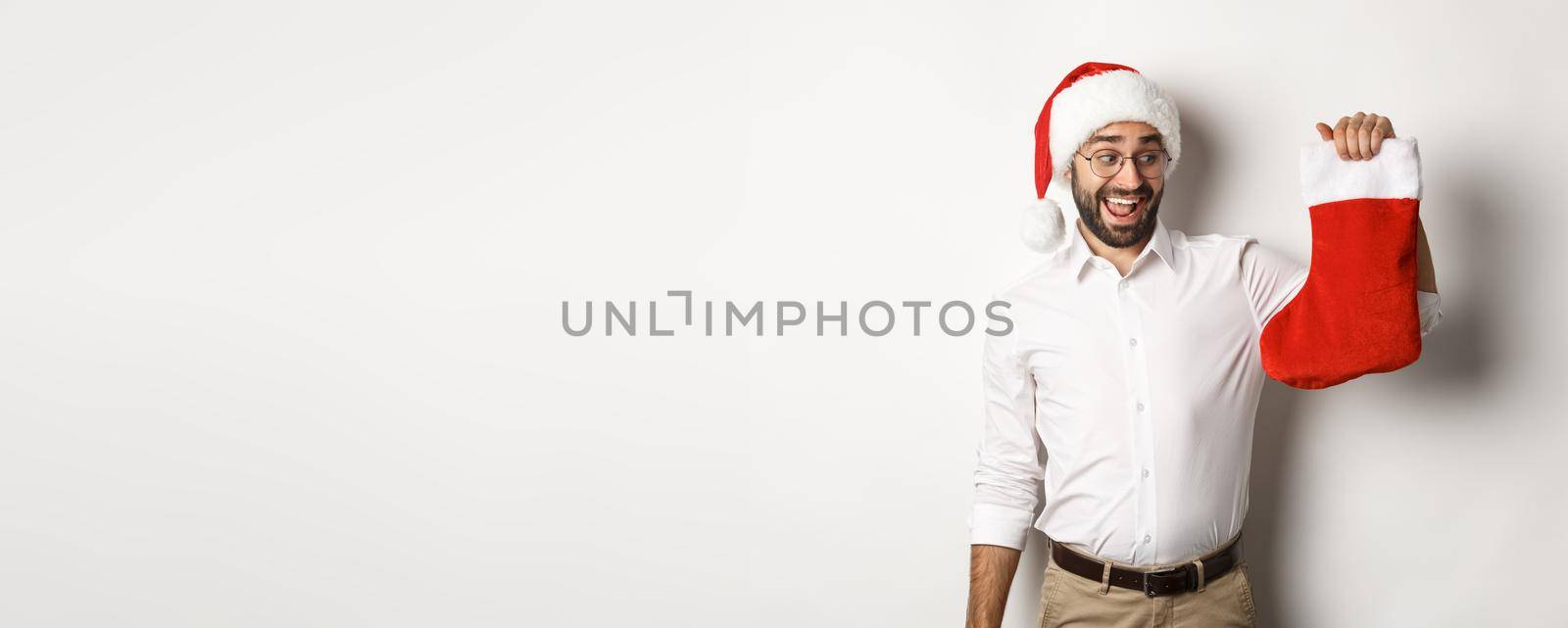 Merry christmas, holidays concept. Happy adult man receive gifts in xmas sock, looking excited, wearing santa hat, white background by Benzoix