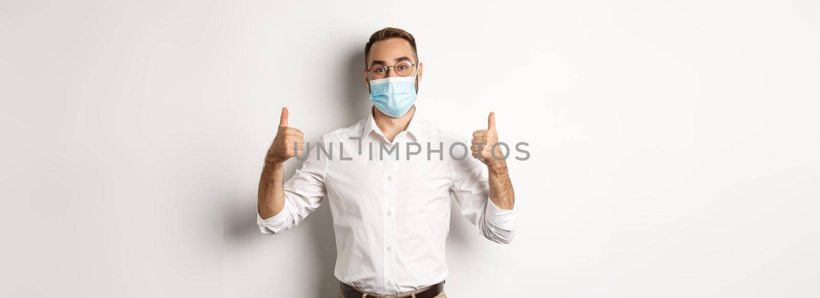 Covid-19, social distancing and quarantine concept. Satisfied male manager showing thumbs up, recommending to wear face mask, standing over white background by Benzoix