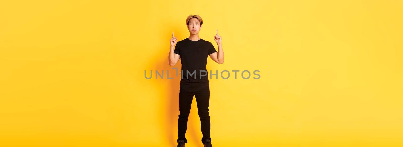 Full-length of sad handsome korean guy with blond hair making upset grimace, pointing fingers up, showing logo with regret, standing yellow background.