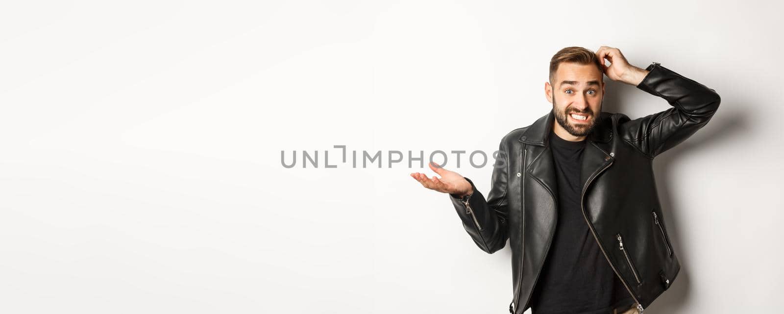 Confused bearded guy in cool leather jacket shrugging, scratch head puzzled and clueless, standing over white background.