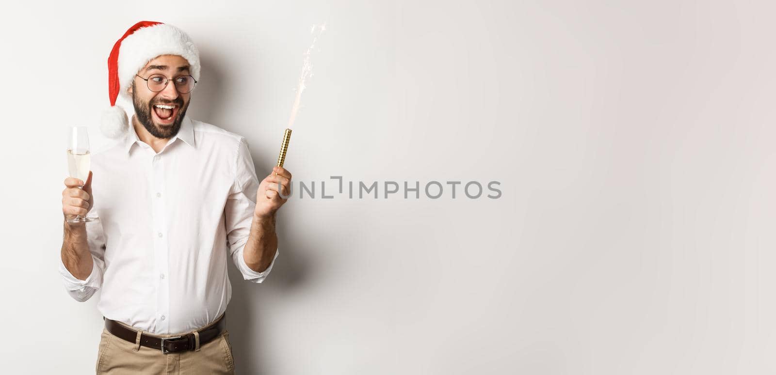 Winter holidays and celebration. Excited man celebrating New Year eve with fireworks sparkles and drinking champagne, wearing santa hat, white background.