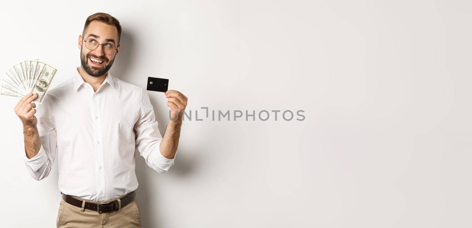 Image of young businessman holding credit card and money, looking at upper left corner and thinking about shopping, standing over white background.