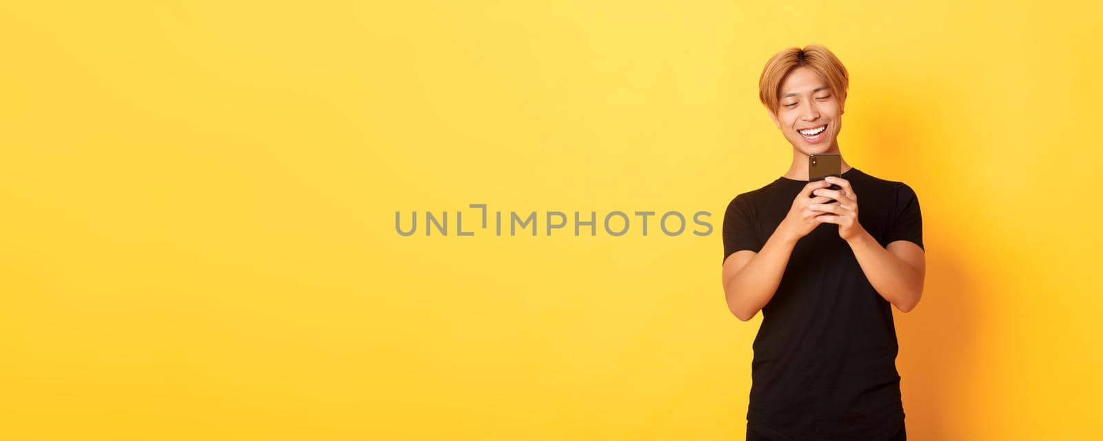 Portrait of handsome stylish asian guy with blond hair, using mobile phone and smiling, messaging in smartphone app, yellow background by Benzoix