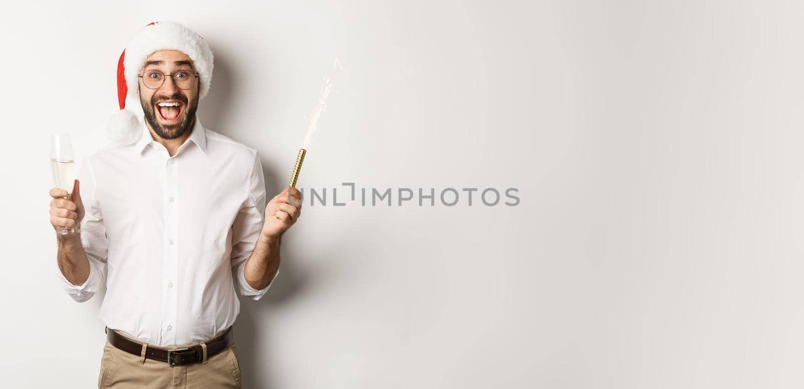 Winter holidays and celebration. Happy guy in Santa hat rejoicing at New Year party, drinking champagne and shouting of joy, white background by Benzoix