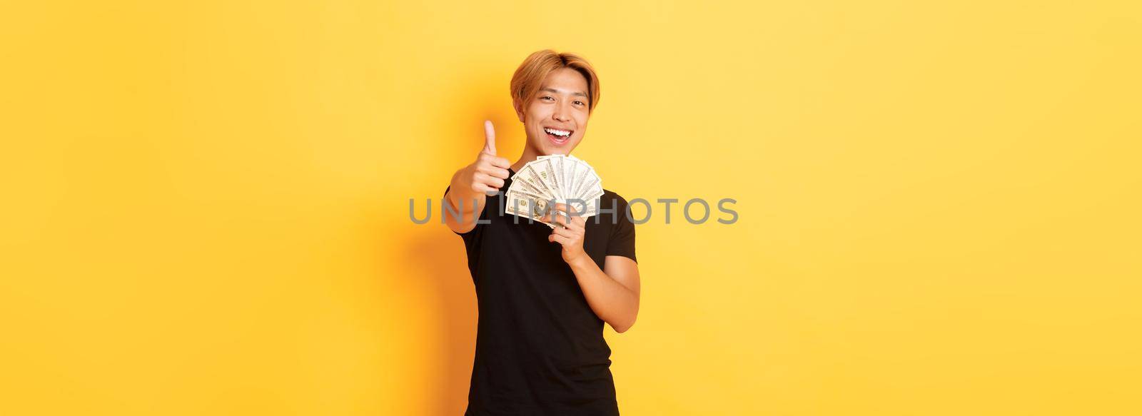 Portrait of handsome confident smiling asian guy showing thumbs-up and holding money, guarantee something, standing yellow background by Benzoix