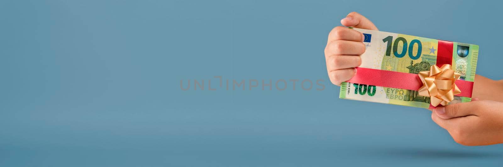 Gift of money. The child holds a stack of banknotes in his hands as a symbol or concept of a gift. 100 euro banknotes tied with a red ribbon with a bow on a blue background