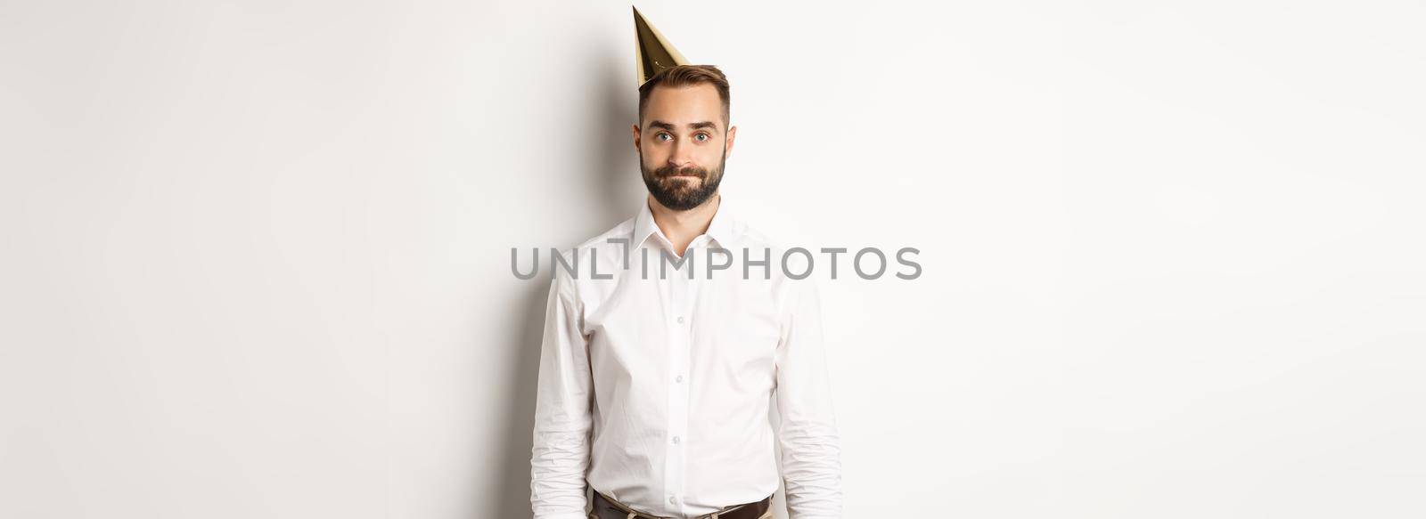 Celebration and holidays. Gloomy guy in birthday hat standing awkward against white background, feeling unamused by Benzoix