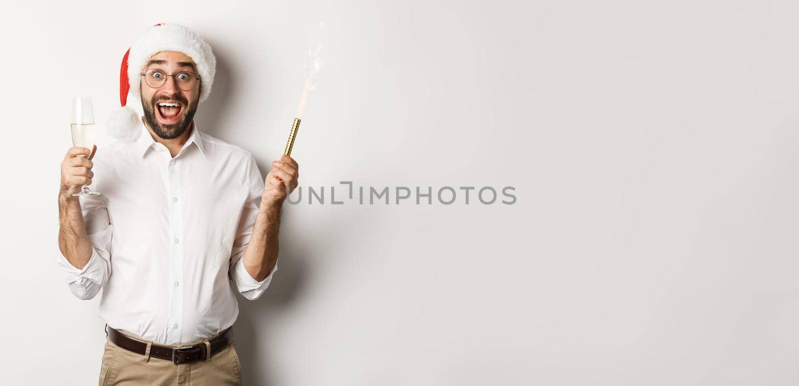 Winter holidays and celebration. Handsome bearded man having New Year party, holding firework sparkler and champagne, wearing santa hat, white background.