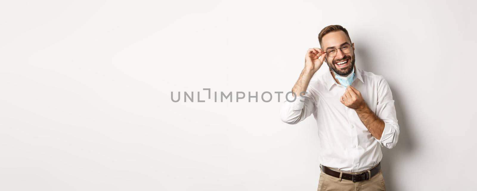 Covid-19, social distancing and quarantine concept. Happy businessman take off face mask and smiling, standing over white background.
