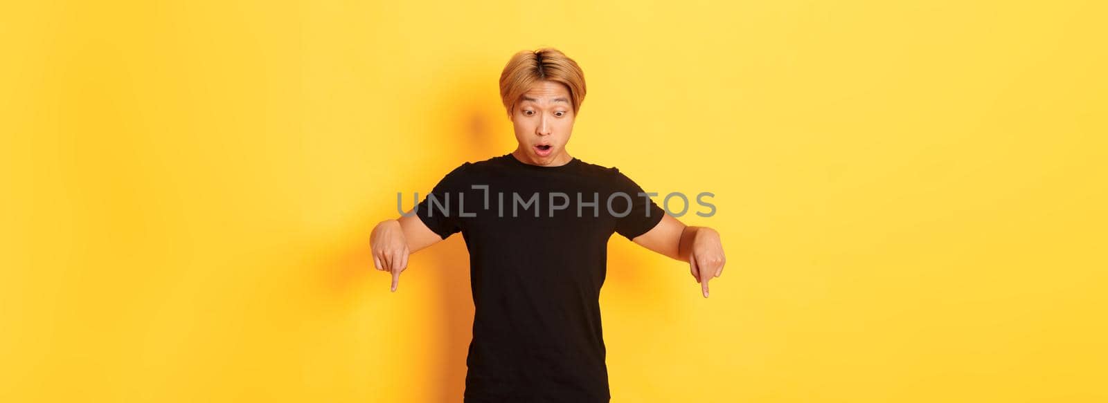 Portrait of surpirsed asian guy gasping amazed, pointing fingers and looking down at banner, standing yellow background by Benzoix