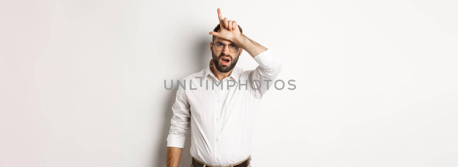 Shocked guy showing loser sign on forehead, complaining, standing over white background by Benzoix