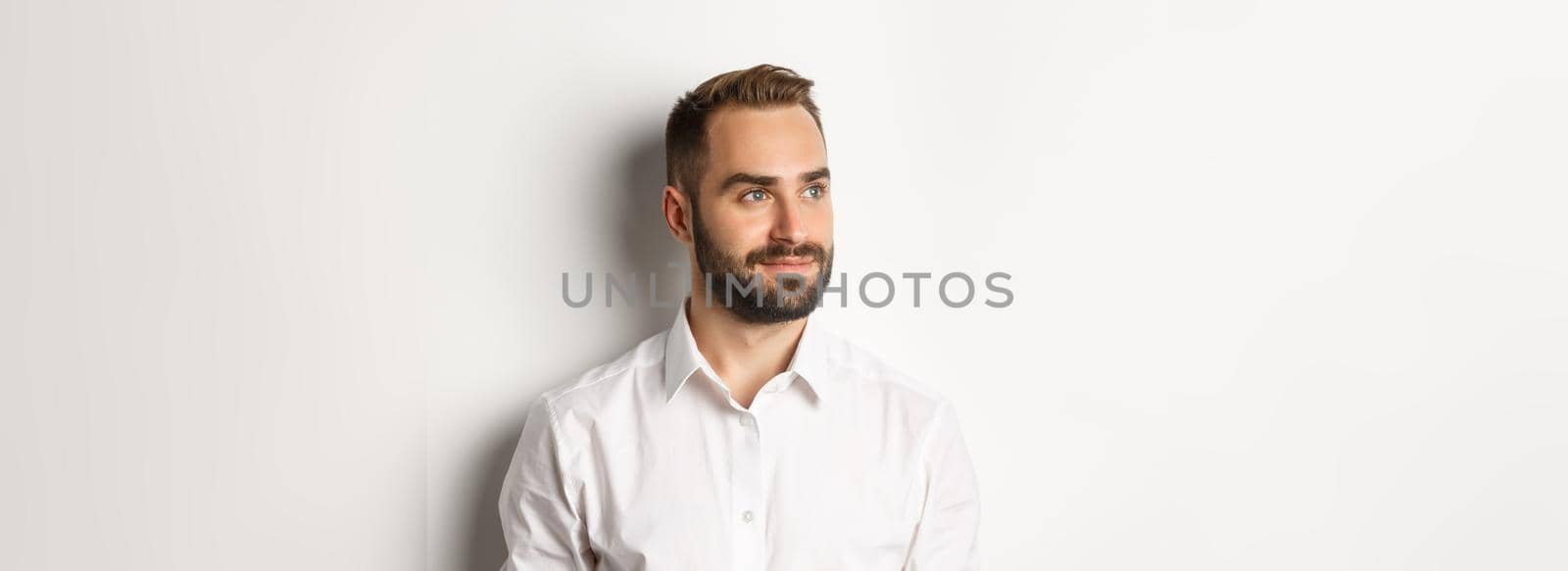 Close-up of confident businessman in white shirt, looking left and smiling satisfied, standing over white background.