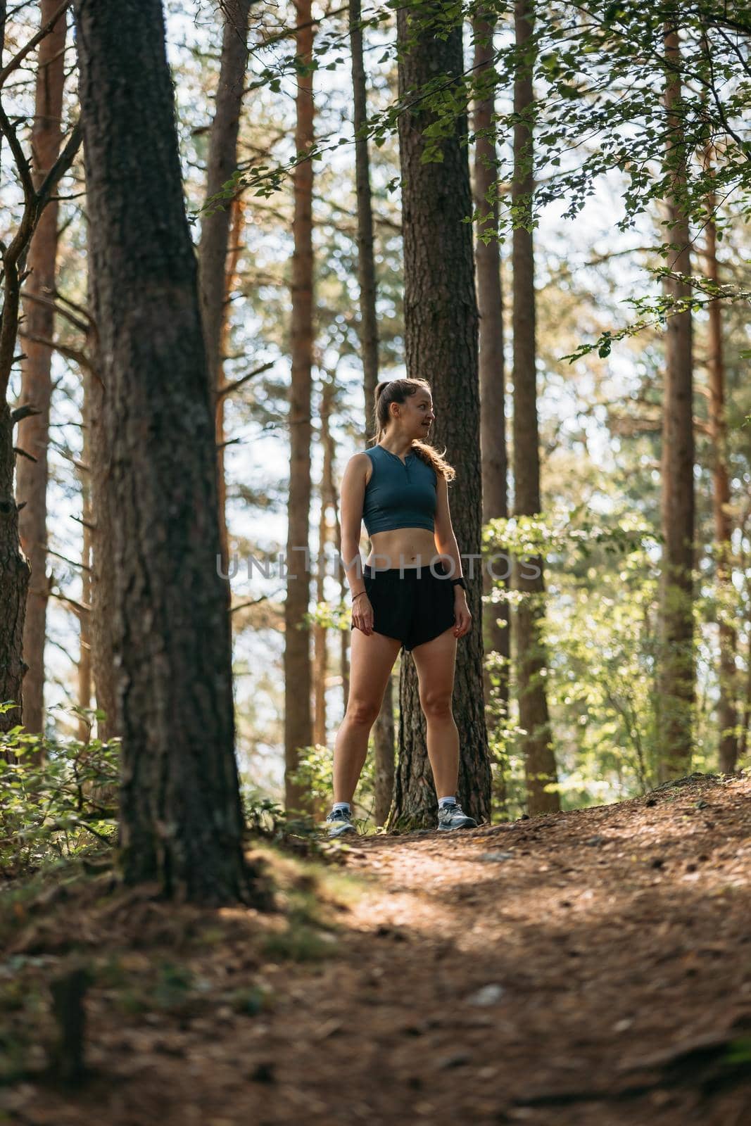 Portrait of young woman runner dressed in sportswear standing on trail in green forest, ready to run while preparing for marathon.