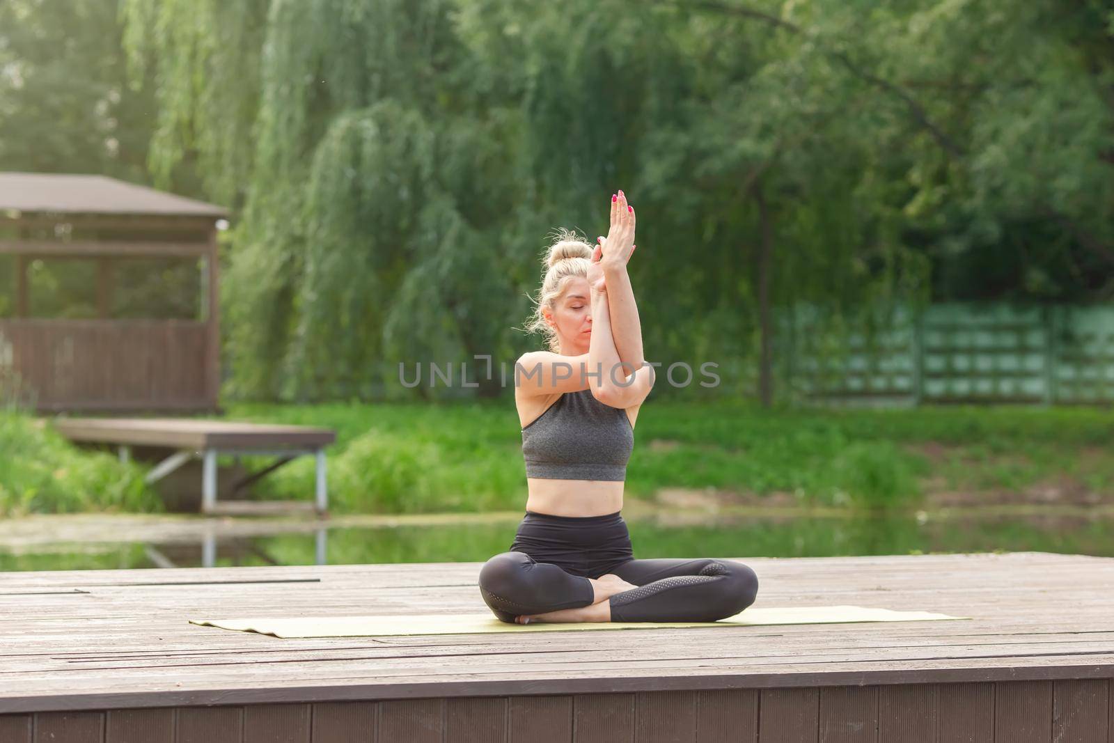 A beautiful woman in a gray top and leggings, sitting on a wooden platform by a pond in the park in summer, does yoga. Copy space.