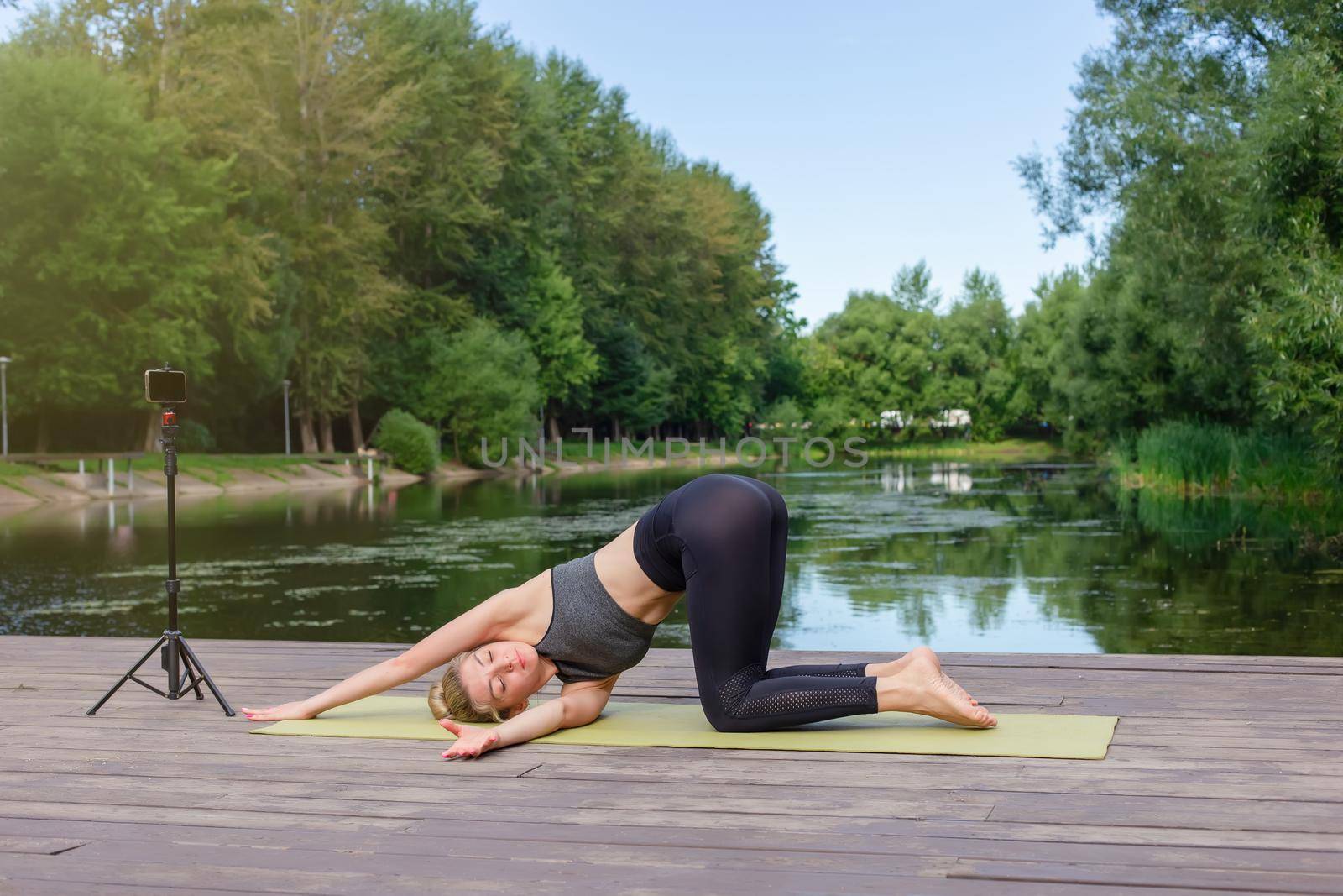 A slender woman in a gray top and leggings, on a wooden platform by a pond in the park in summer, does yoga, performs twisting exercises, on a green sports mat, next to her smartphone on a tripod.