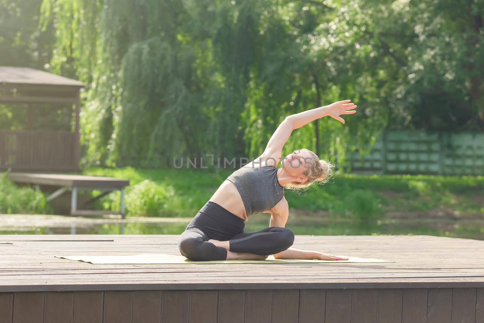 A slender woman in a gray top and leggings, sitting on a wooden platform by a pond in the park in summer and performing a body tilt sideways with her arm raised. Copy space
