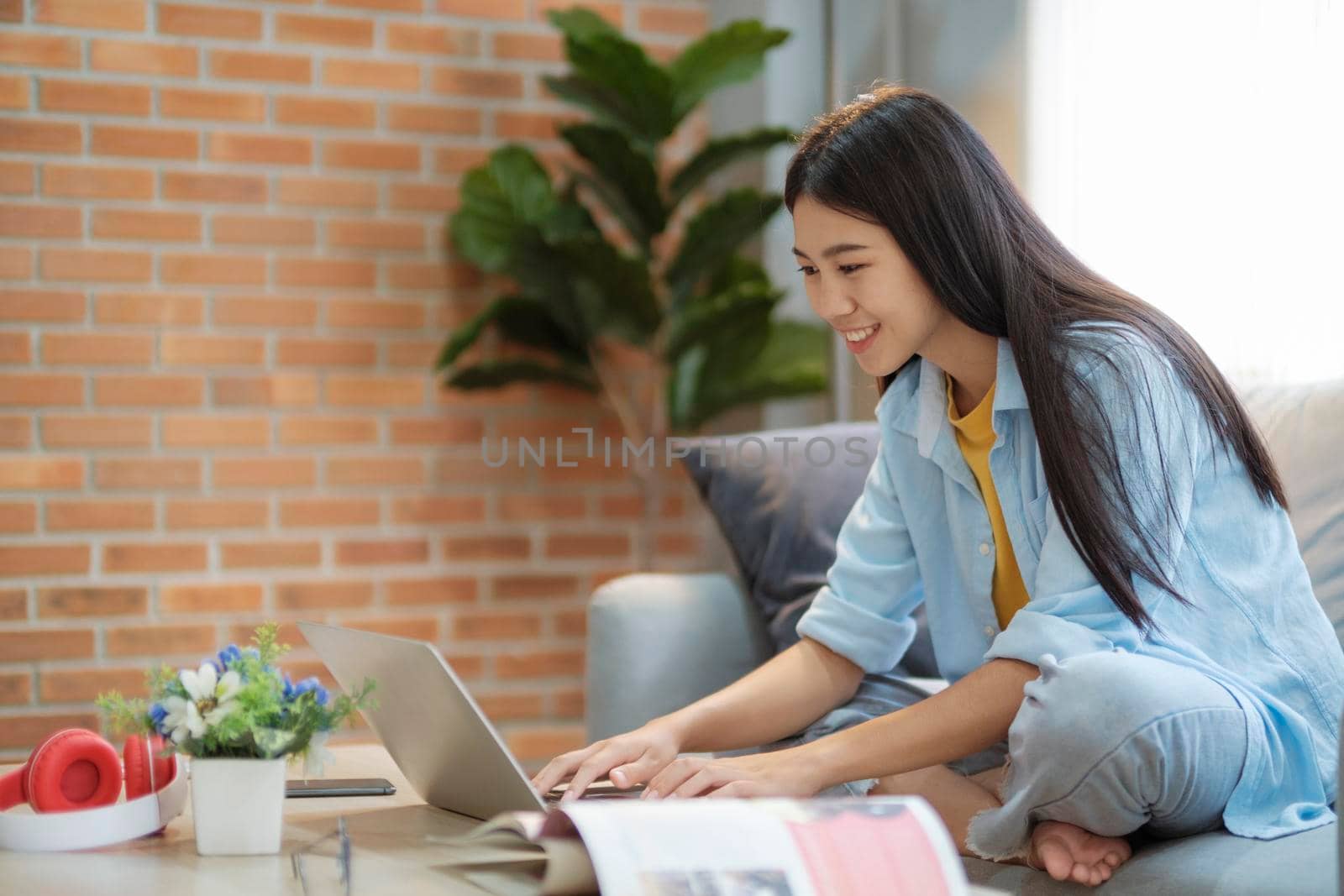 Young asian woman surfing the internet using laptop and holding book while sitting on couch at home. Young asian women studying online at home. Online connection and online learning concept.