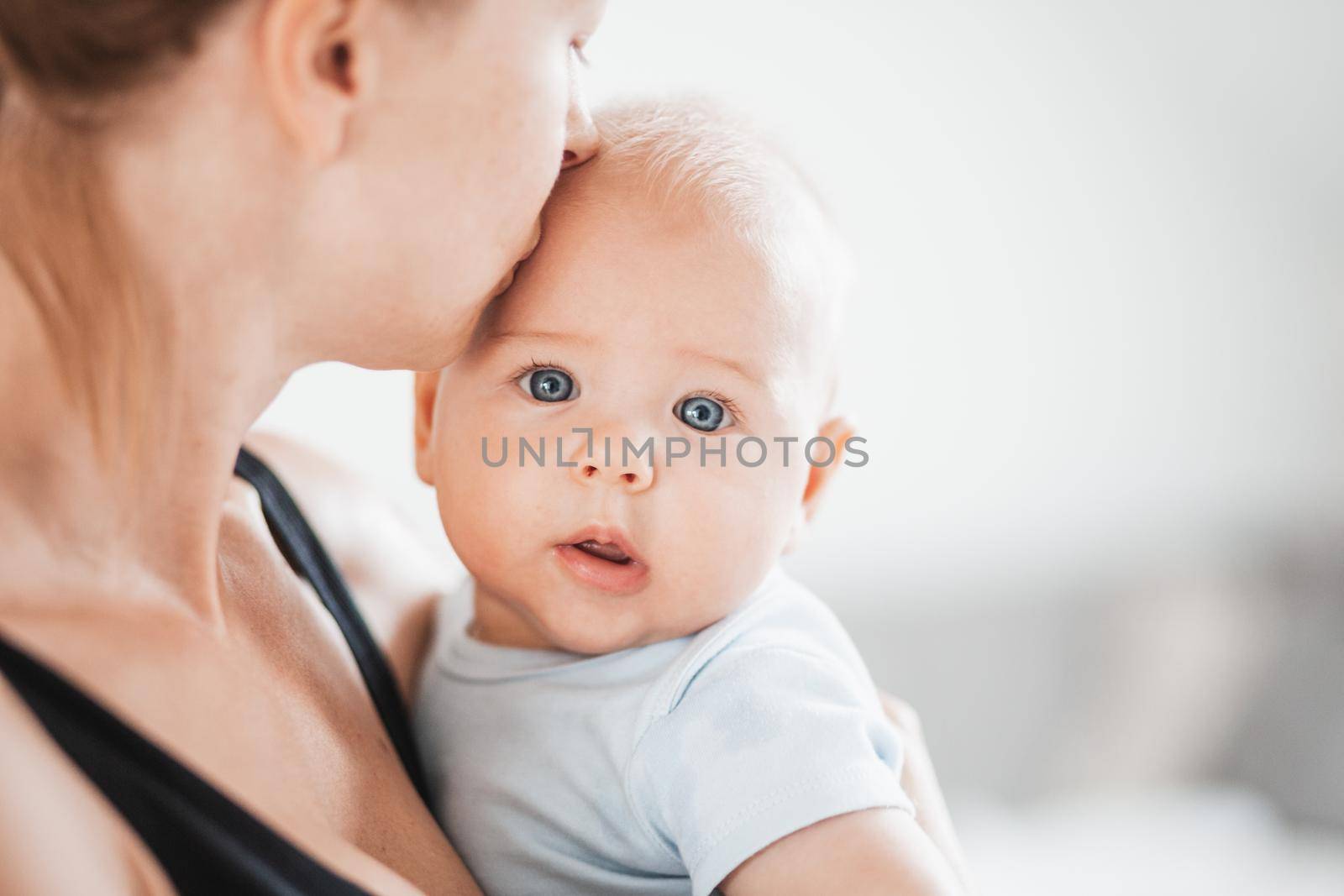 Portrait of sweet baby resting in mothers arms, looking at camera. New mom holding and kissing little kid, embracing child with tenderness, love, care. Motherhood concept.