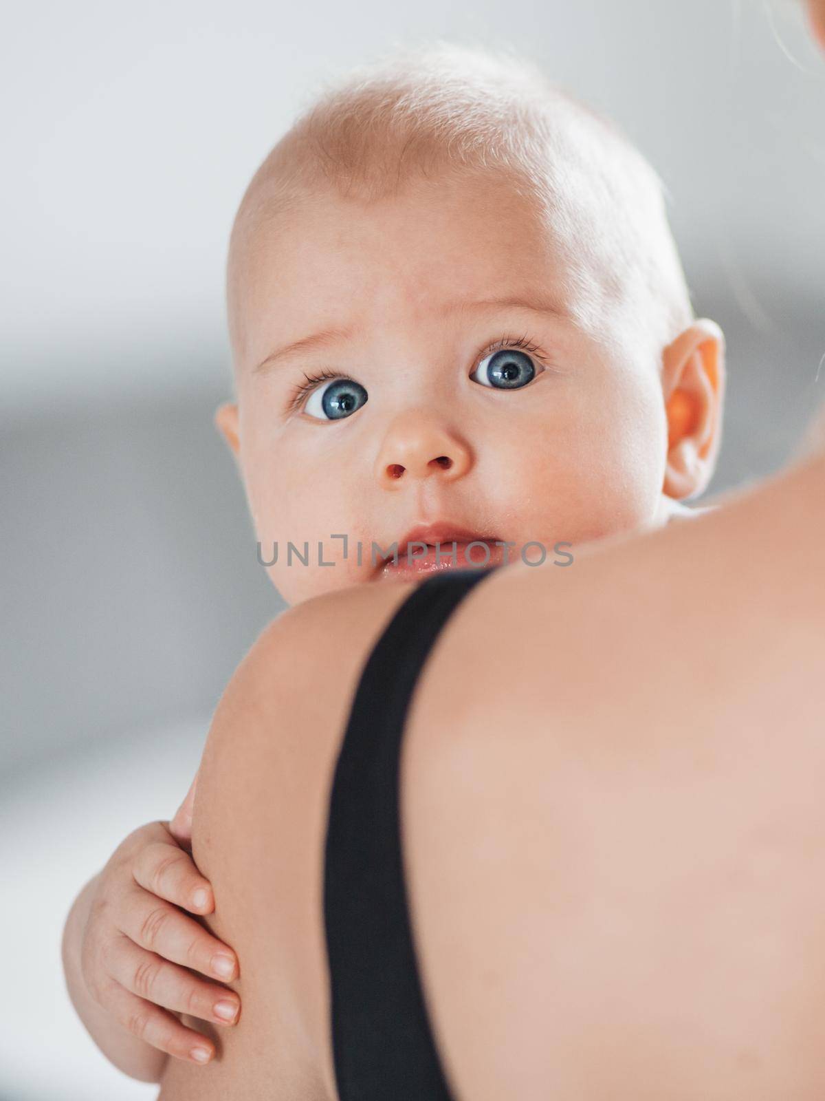 Portrait of sweet baby resting in mothers arms, looking at camera, touching mama shoulder. New mom holding little kid, embracing child with tenderness, love, care. Motherhood concept.