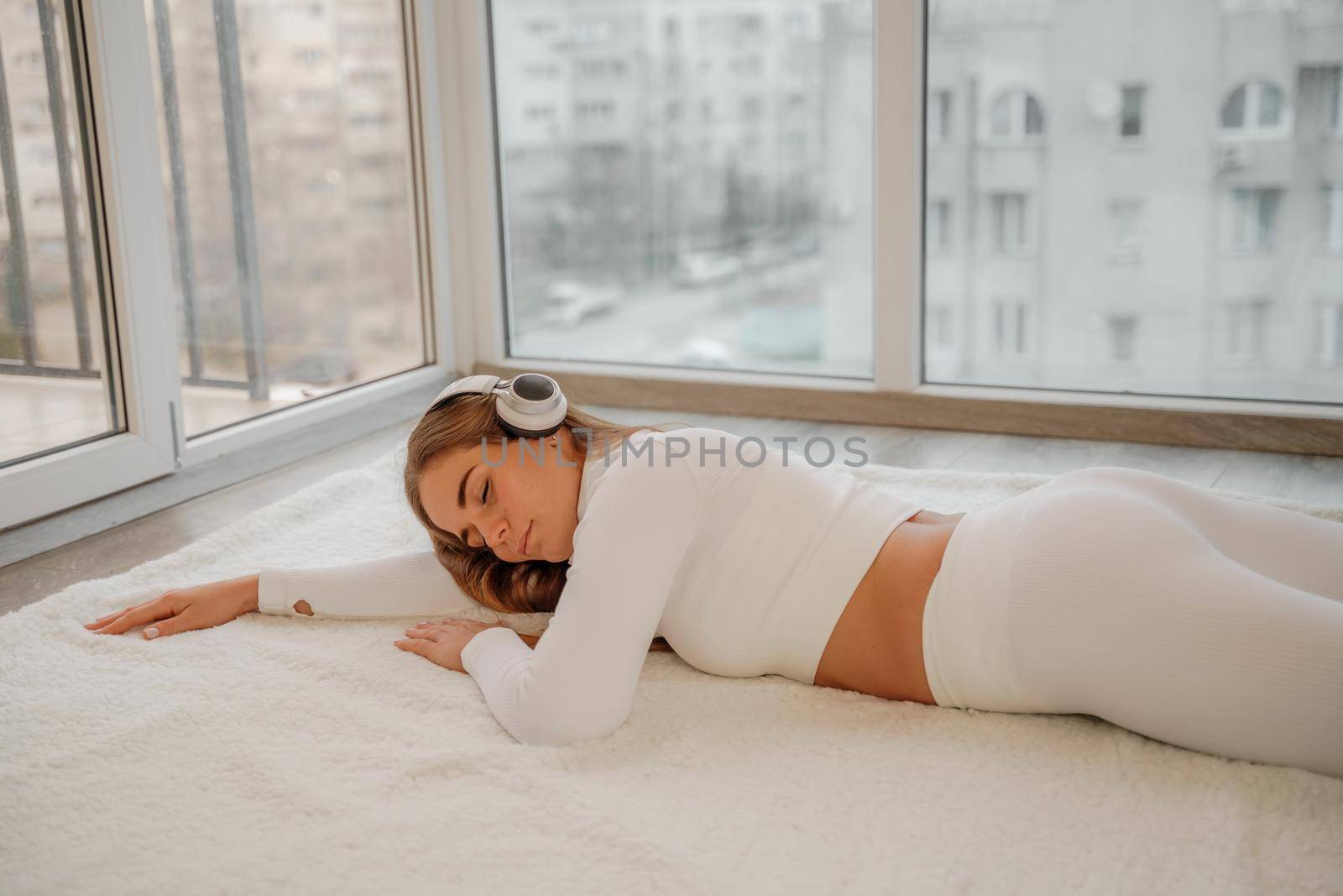 Side view portrait of relaxed woman listening to music with headphones lying on carpet at home. She is dressed in a white tracksuit