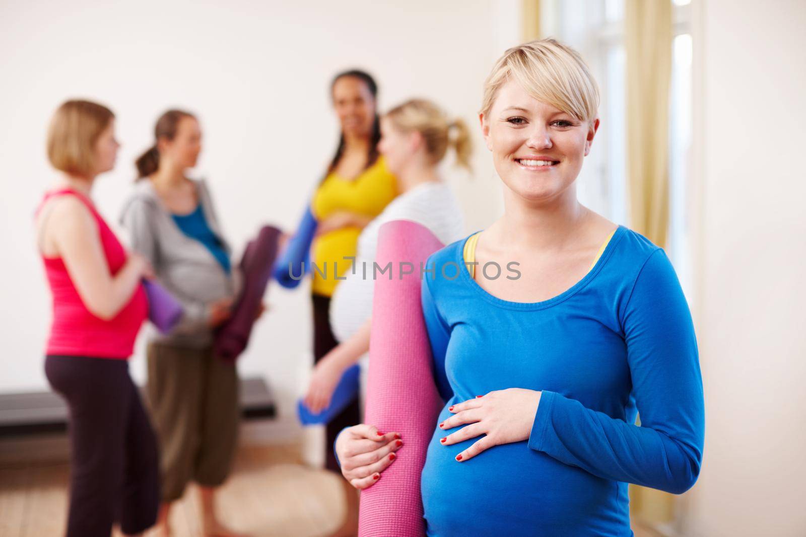 Pilates for pregnancy. A young blonde pregnant woman in a gym holding an exercise mat with a group of women in the background. by YuriArcurs