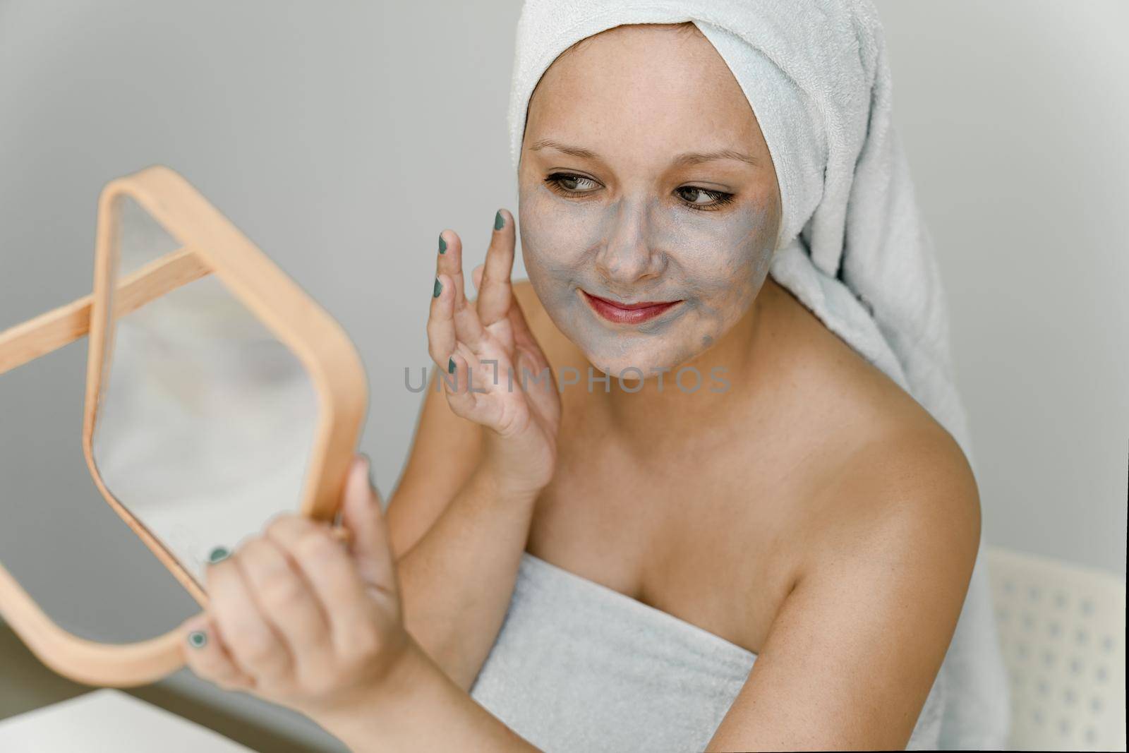 Young woman puts gray cosmetic clay on her face while looking in mirror, her hair and body are wrapped in towel. Close-up