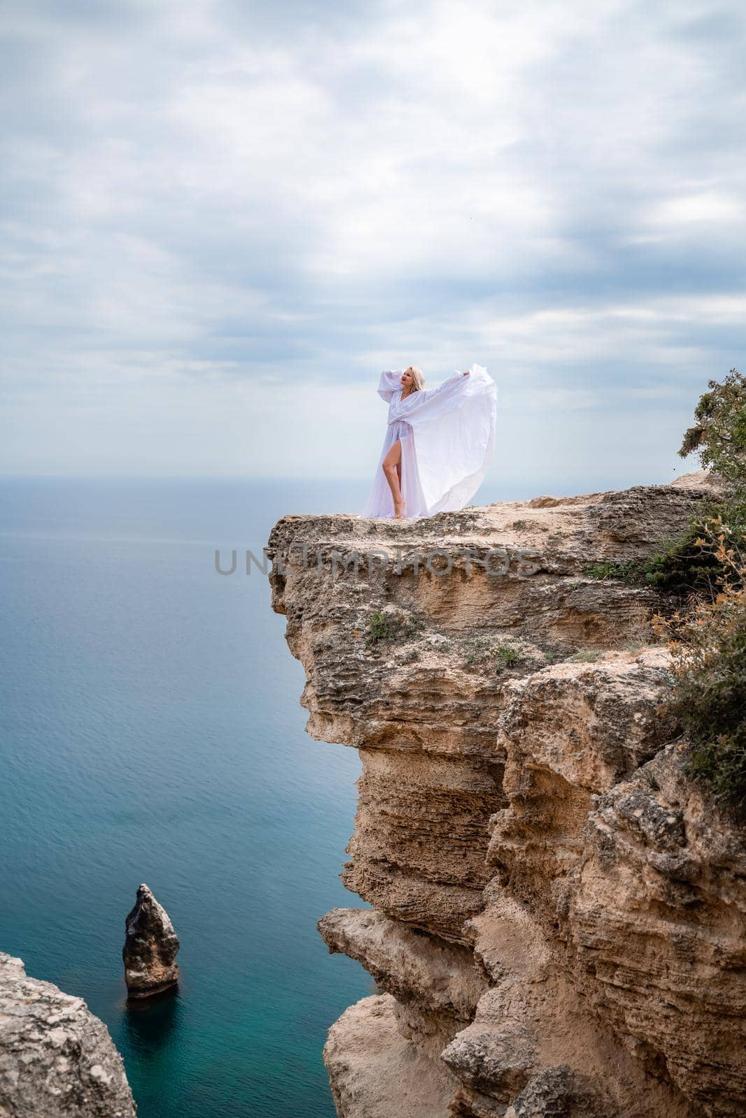 Blonde with long hair on a sunny seashore in a white flowing dress, rear view, silk fabric waving in the wind. Against the backdrop of the blue sky and mountains on the seashore. by Matiunina