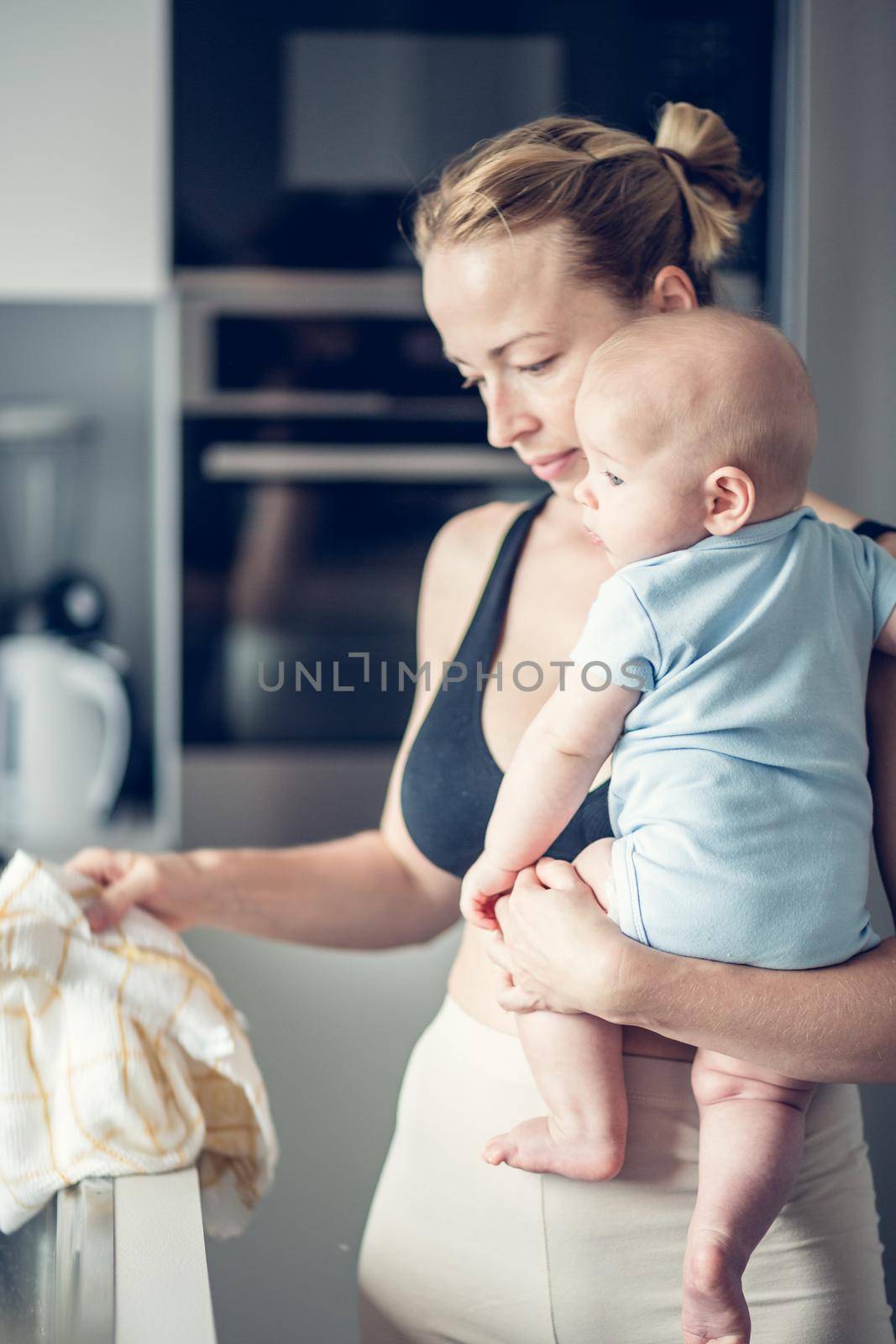 Woman wiping kitchen sink with a cloth after finishing washing the dishes while holding four months old baby boy in her hands.