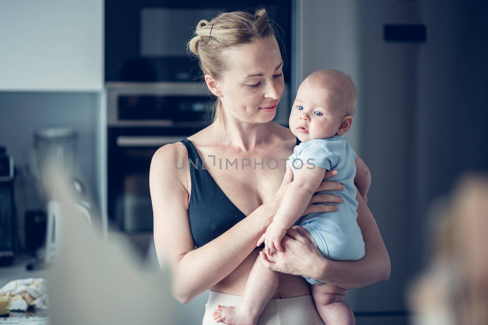 Pretty young mother holding her newborn baby boy standing near kitchen window at home.