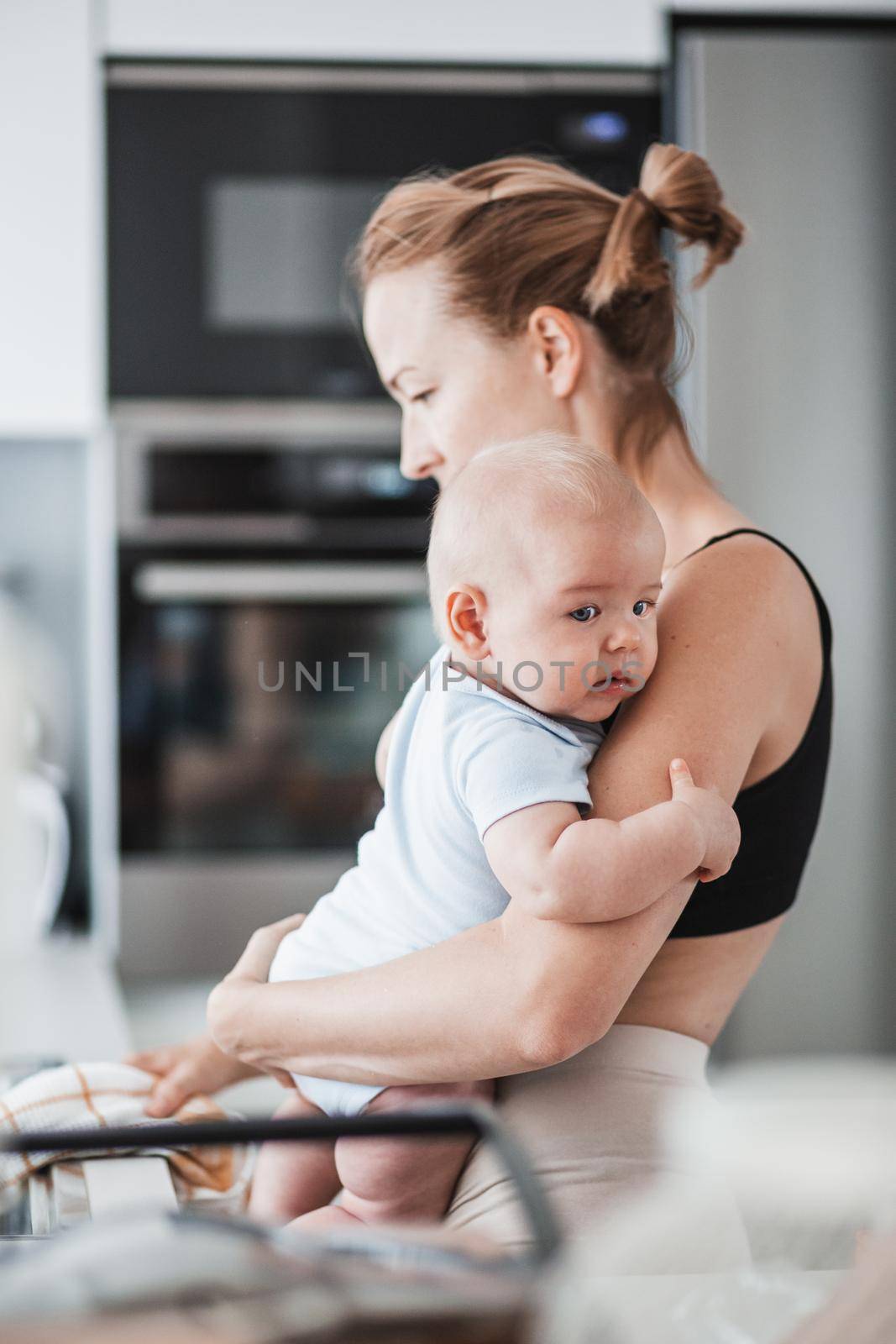 Woman wiping kitchen sink with a cloth after finishing washing the dishes while holding four months old baby boy in her hands by kasto