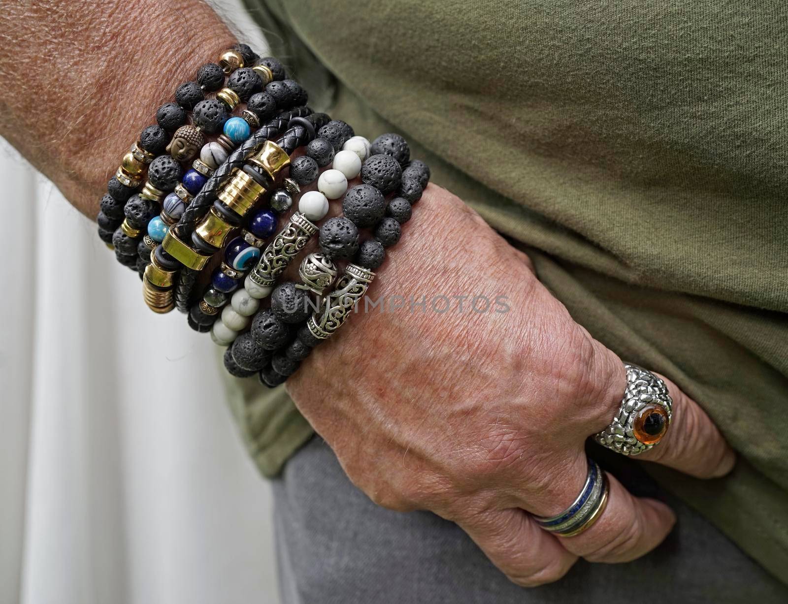 Senior man putting his hand in the pocket of his grey jeans. He wears lava stone and other breaded bracelets and special rings.
