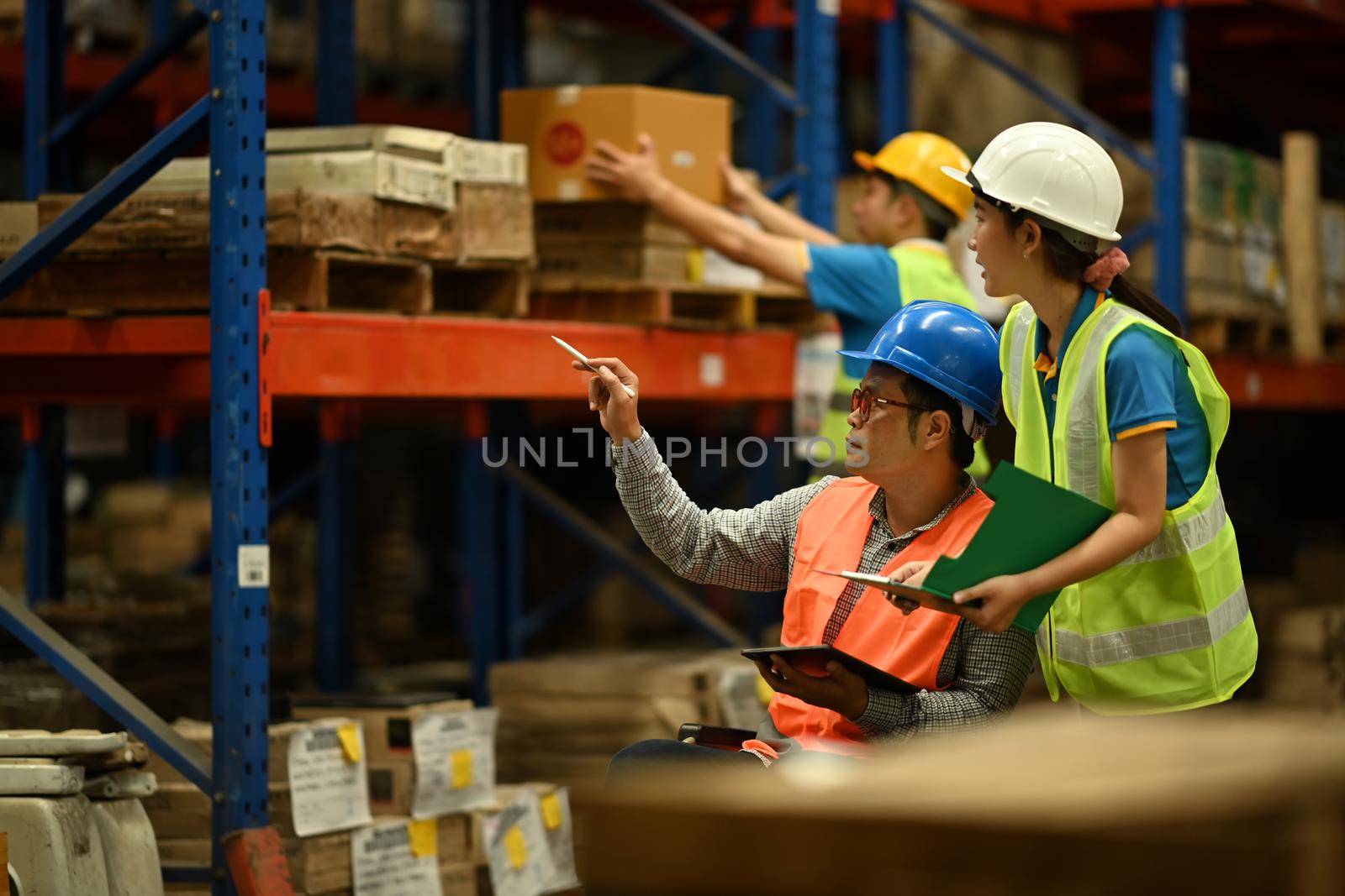 Senior male manager in wheelchair and young workers inspecting stock tick and cardboard stock product in a large warehouse by prathanchorruangsak