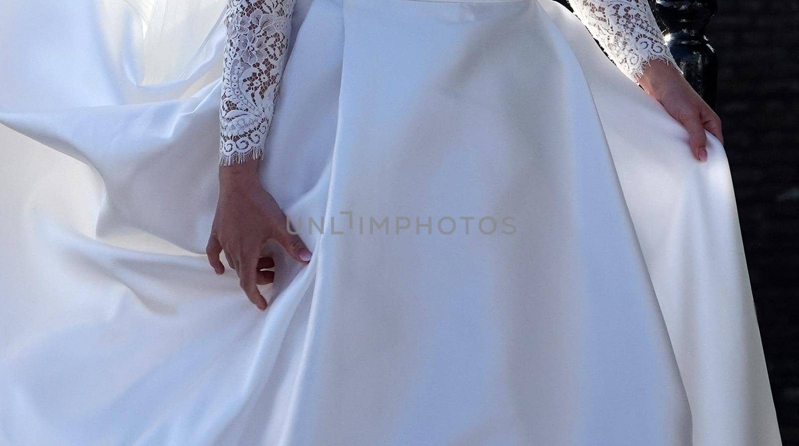 The hands of a bride. She lifts her long skirt to walk. The sleeves of her white dress are made of lace.