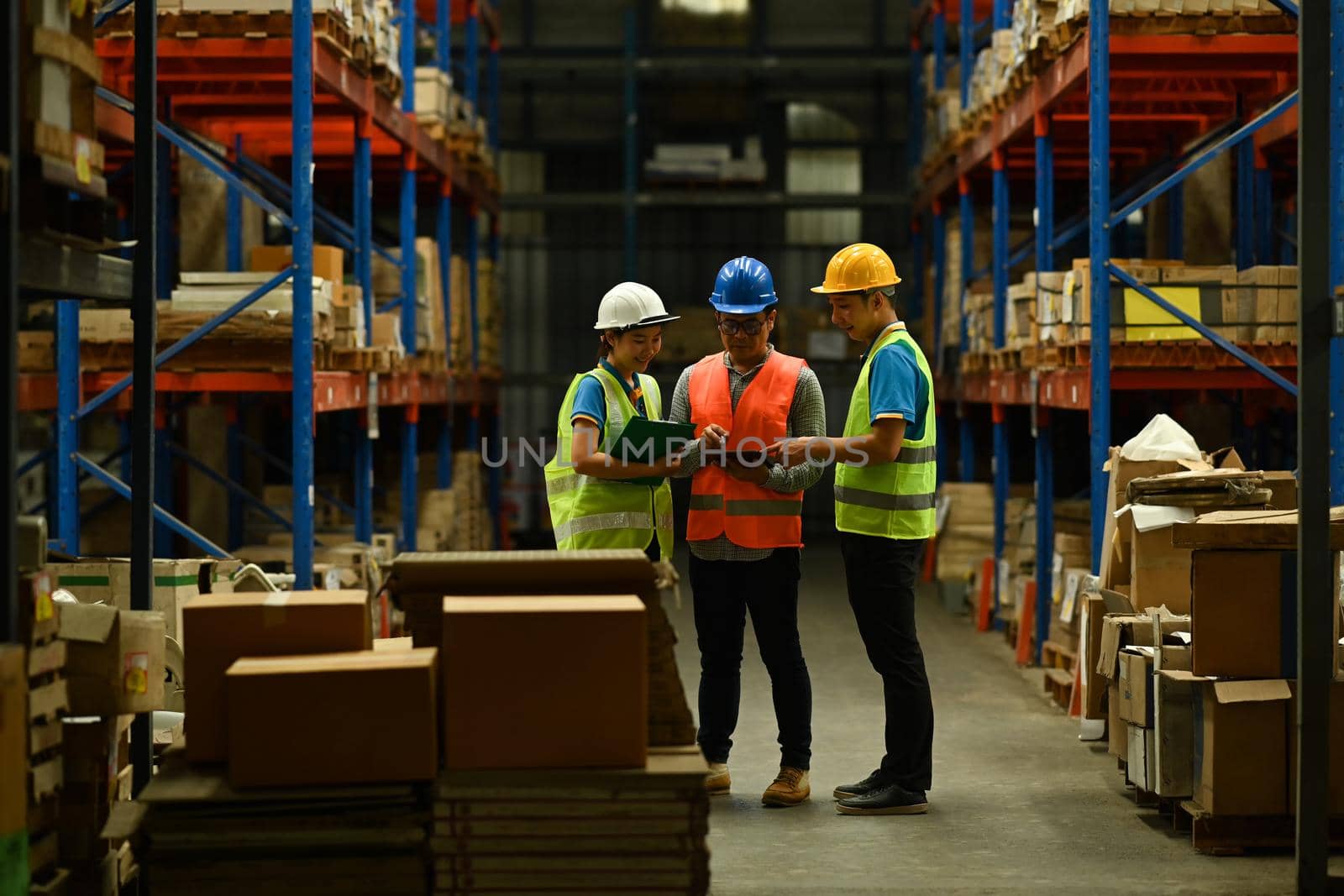 Senior male managers and storehouse employees standing between rows of tall shelves and checking inventory on digital tablet by prathanchorruangsak