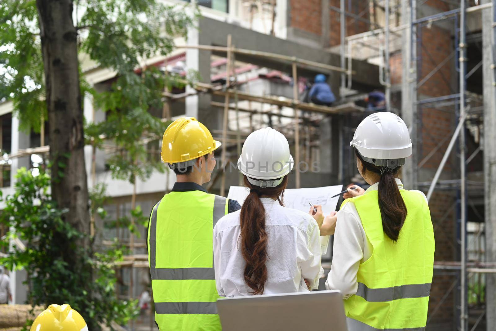 Rear view of inspectors team in safety helmet working with blueprints and supervising progress of industrial building construction project.