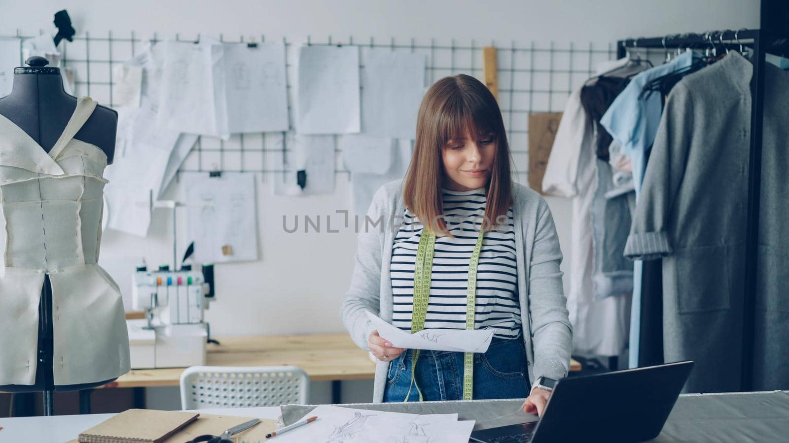 Young attractive woman is choosing sketches for new fashion show and using laptop in her modern tailor's shop. Creative thinking in clothing design industry concept.