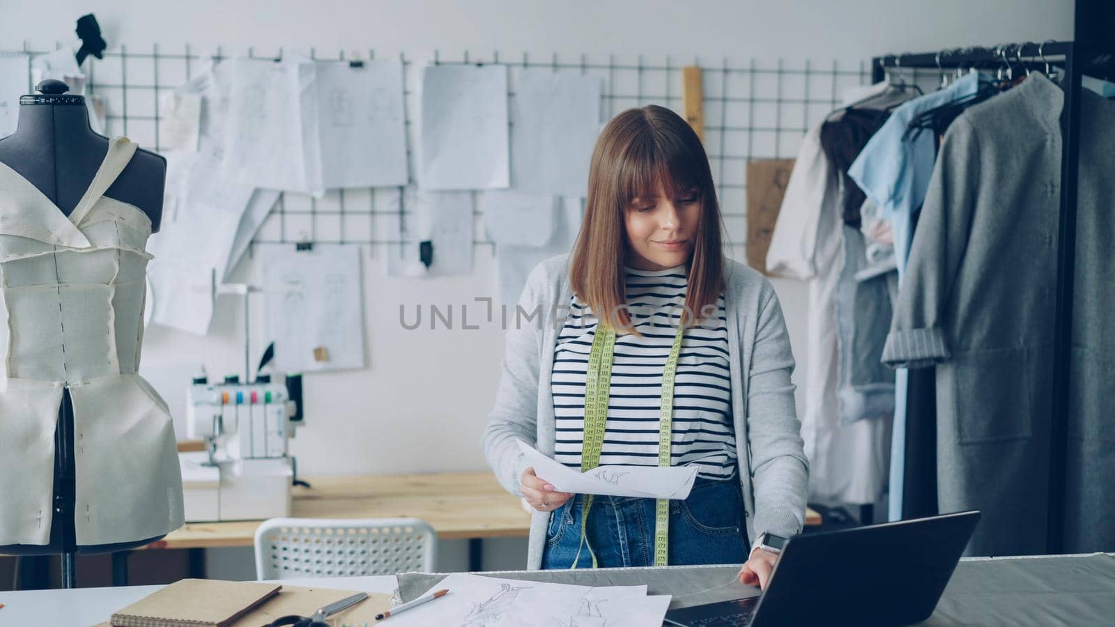 Young attractive woman is choosing sketches for new fashion show and using laptop in her tailor's shop. Creative thinking in clothing design industry concept. by silverkblack