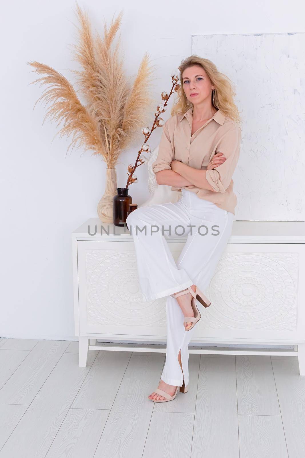 a beautiful slender woman, 30-40 years old, with blond hair, sits in a beige shirt and white trousers in the interior of a white room, against a wall and a chest of drawers. Copy space. Vertical