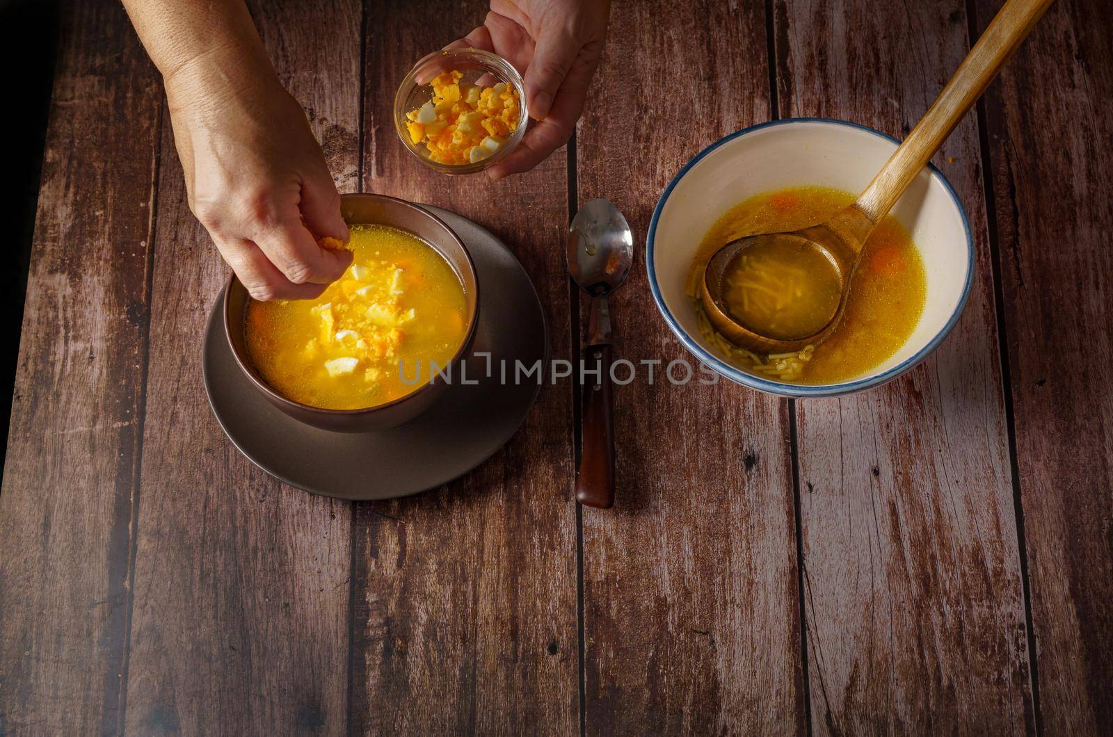 woman pouring hard-boiled egg into a chicken noodle soup in a brown bowl on a wooden table and a white soup tureen