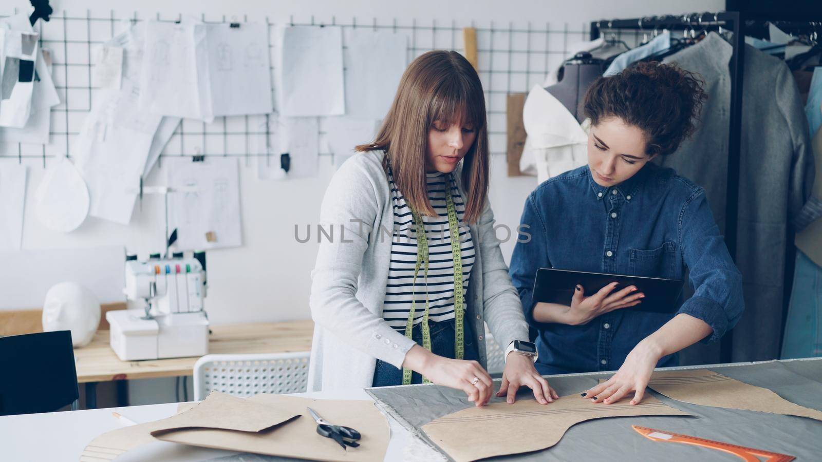 Young female tailors are busy working with tablet, communicating and outlining clothing pattern on textile on studio desk. Light workshop with sewing items in background.