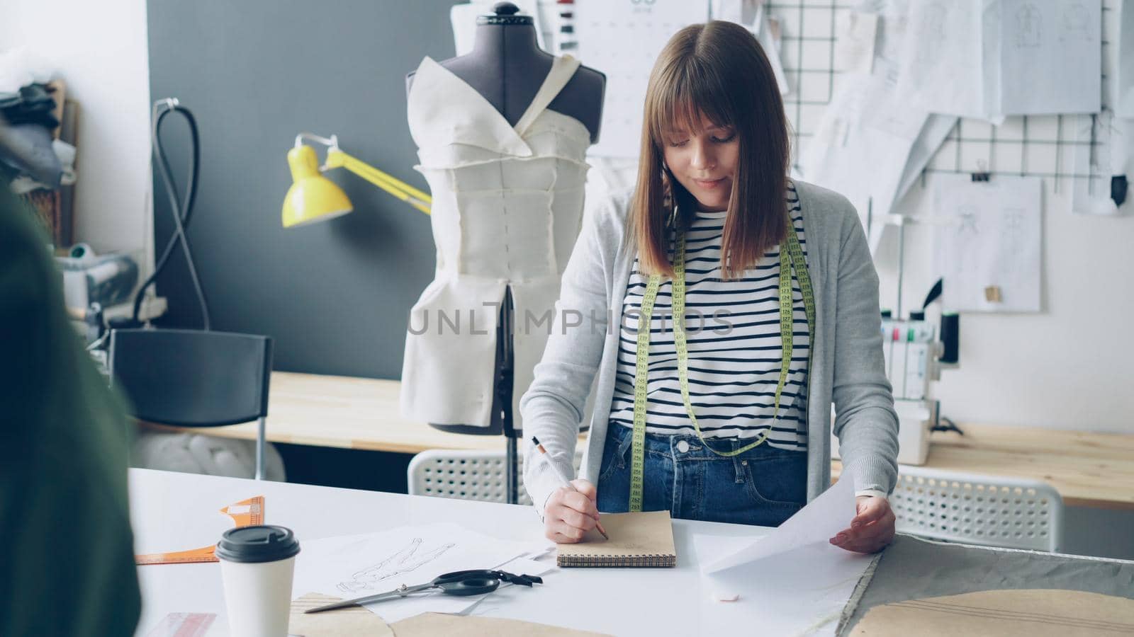 Young woman is drawing in notepad looking at clothing sketch while working in her tailoring studio. Drawings hanging on wall, bright sewing items are visible. by silverkblack