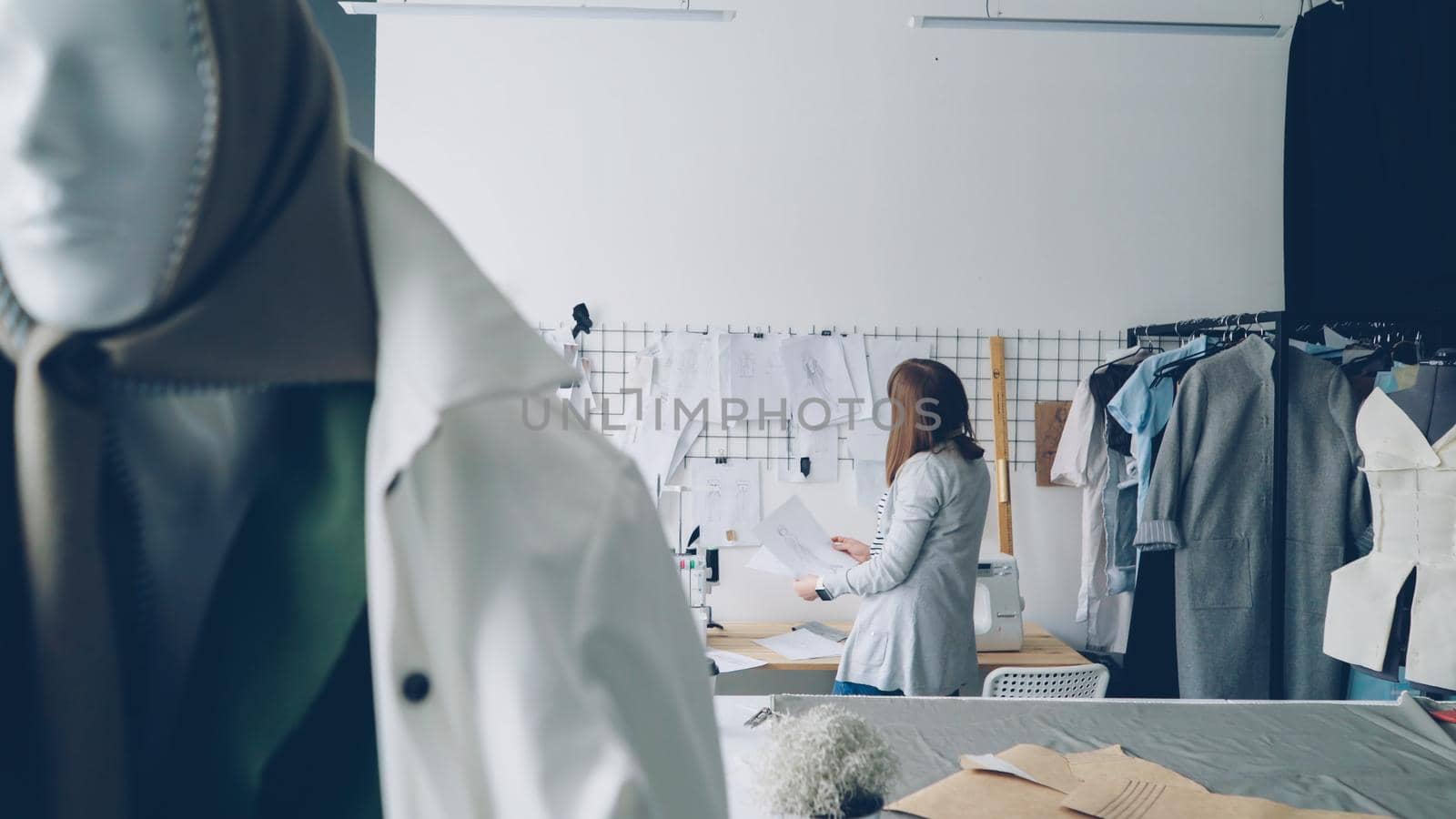 Attractive young woman clothing designer is looking at garment sketches hanging on wall then choosing new drawings. Mannequin dresses in trendy clothes is in foreground.