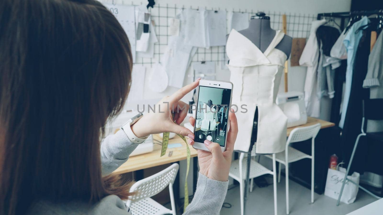 Young female clothing design blogger is shooting tailoring dummy with half-finished garment pinned to it. Close-up shot of girl's hands and smartphone. by silverkblack