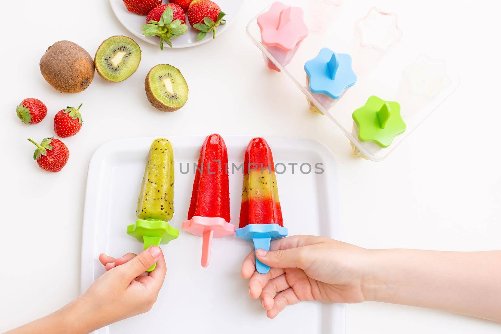 Two childrens hands, take multi-colored homemade kiwi and strawberry ice cream from a white square plate, on a white table, top view