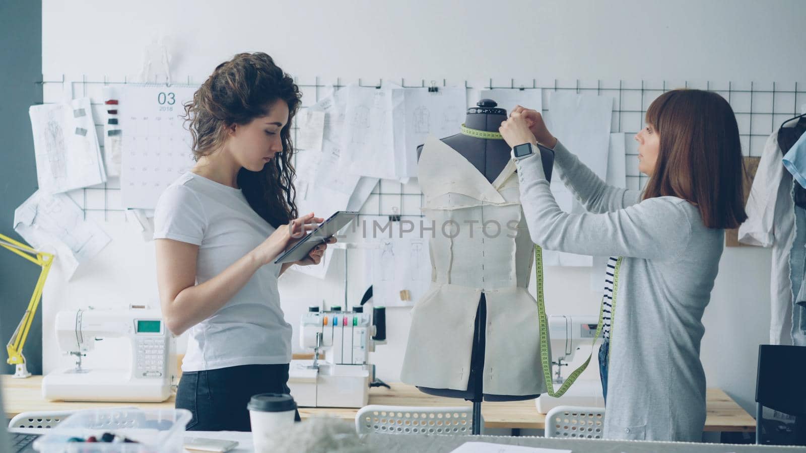 Clothing designer entrepreneurs are measuring mannequin parts while working in nice modern studio. Young attractive woman is using tablet to enter measurement results. by silverkblack