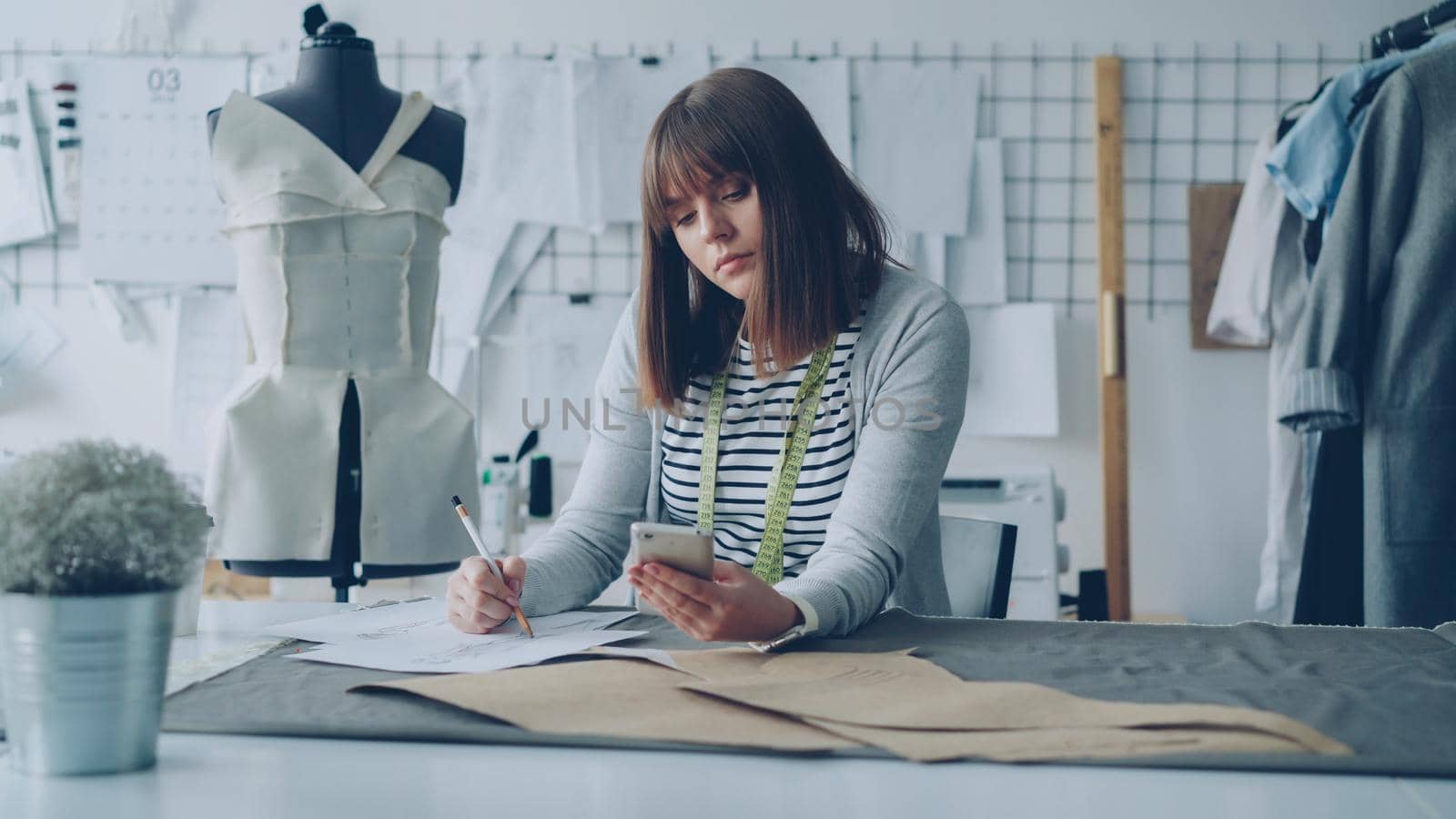 Creative clothing designer is looking at smartphone screen and drawing sketch while working in modern tailor shop at sewing table. Woman is busy and involved in process.